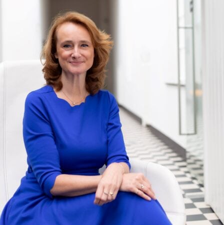 Een vrouw met schouderlang kastanjebruin haar en een blauwe jurk zit op een witte stoel in een lichte, moderne hal met geruite vloer.