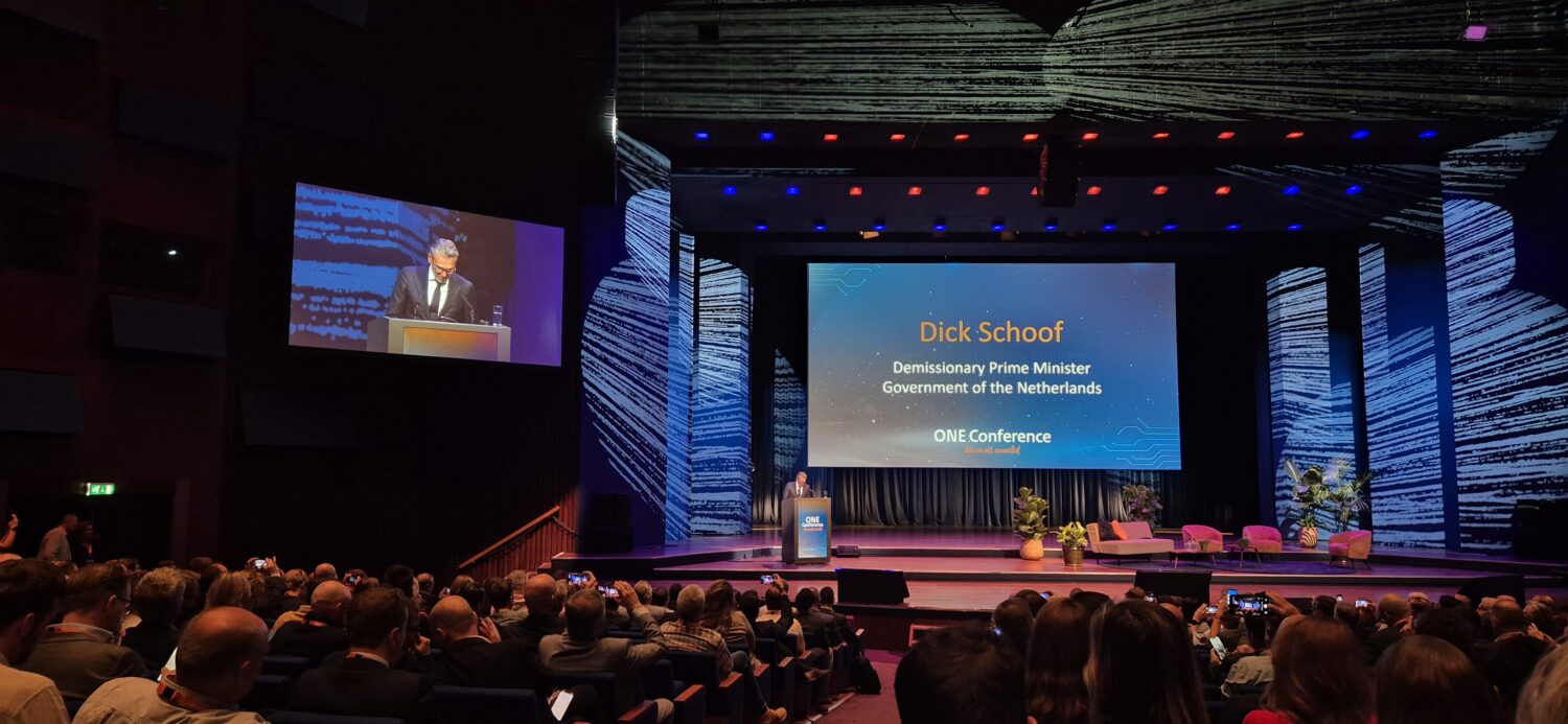 Een spreker op een podium spreekt een publiek in een groot auditorium toe; een scherm erachter toont "Dick Schoof, Demissionair Minister-President, Regering van Nederland, ONE Conferentie.".
