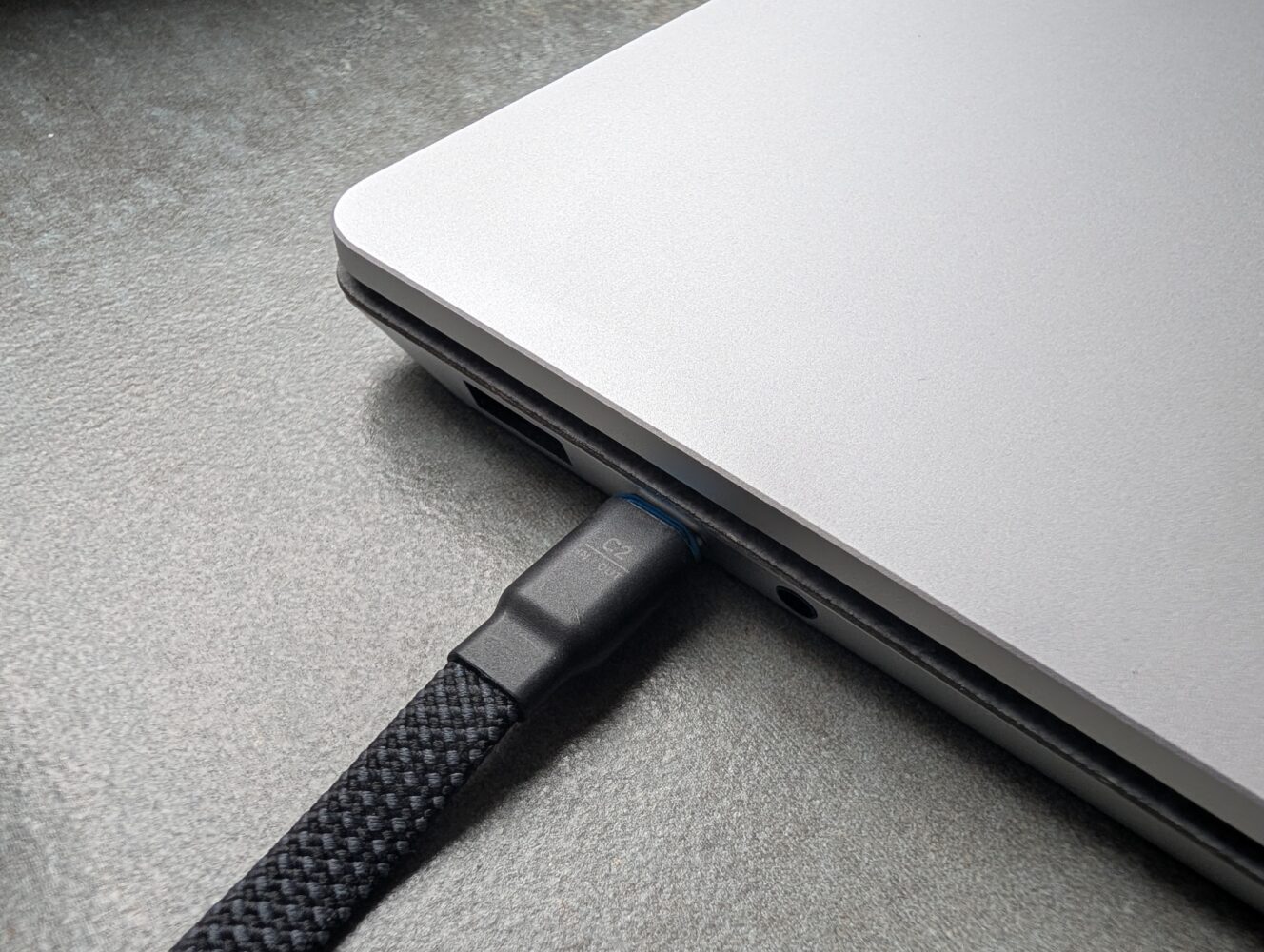 Een gevlochten USB-C kabel verbindt een laptop powerbank met de zijpoort van een gesloten zilveren laptop die op een grijs oppervlak rust.