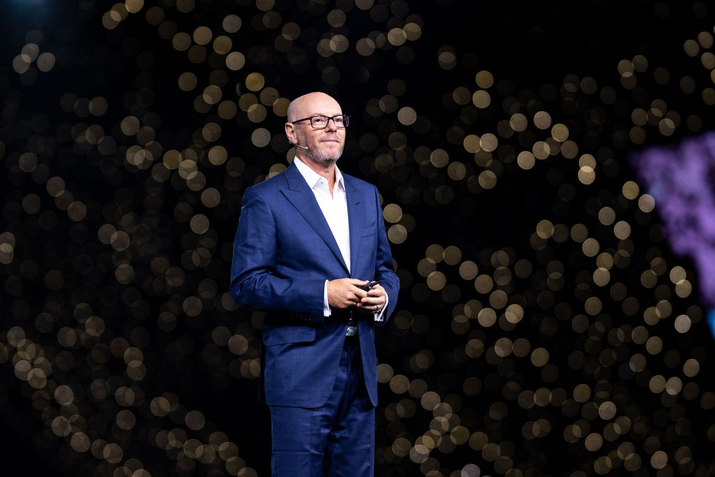 2025_SuiteWorld_ExecutiveKeynote01_88144 (1) Een man in een blauw pak en een bril staat op het podium en houdt een klein voorwerp vast, met vage lichten op de donkere achtergrond.