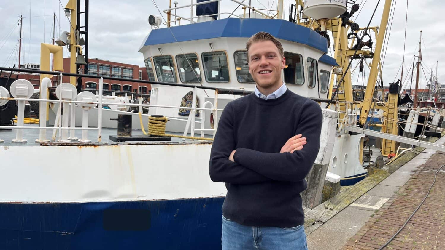 Een man staat met gekruiste armen voor een aangemeerde vissersboot op een bewolkte dag.
