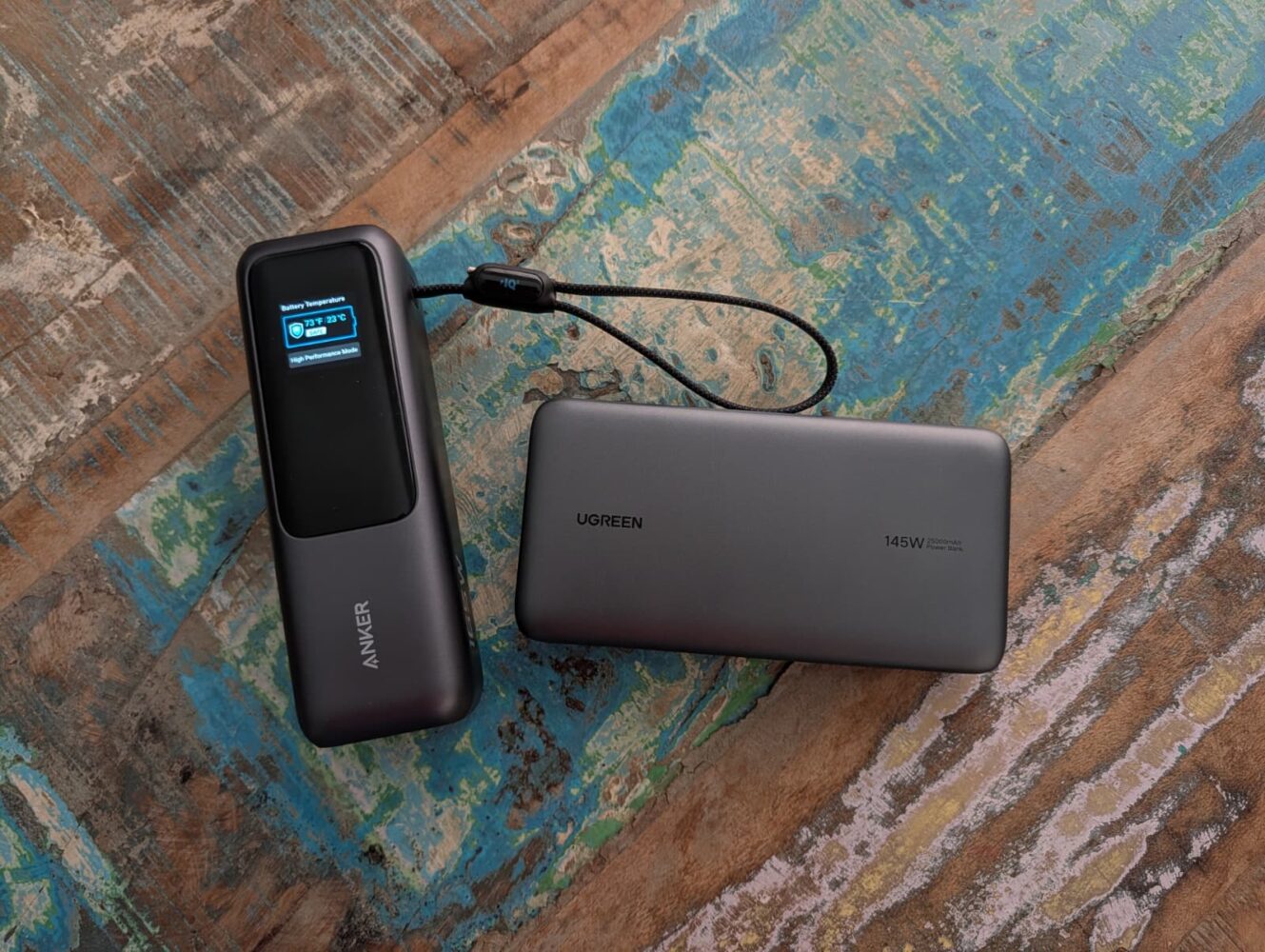 Een Anker Powerbank met display en een Ugreen 145W Powerbank zijn verbonden door een kabel en rusten op een houten oppervlak met textuur.