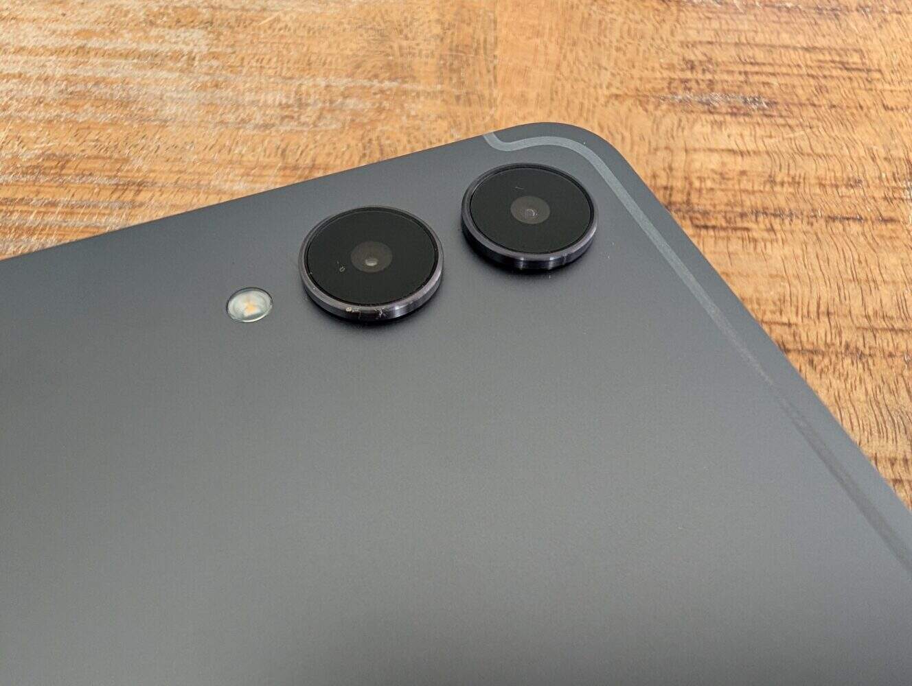 Close-up van de hoek van een Samsung Galaxy Tab S11 Ultra met twee cameralenzen, een kleine flitser en een deel van een matte achterkant op een houten oppervlak.