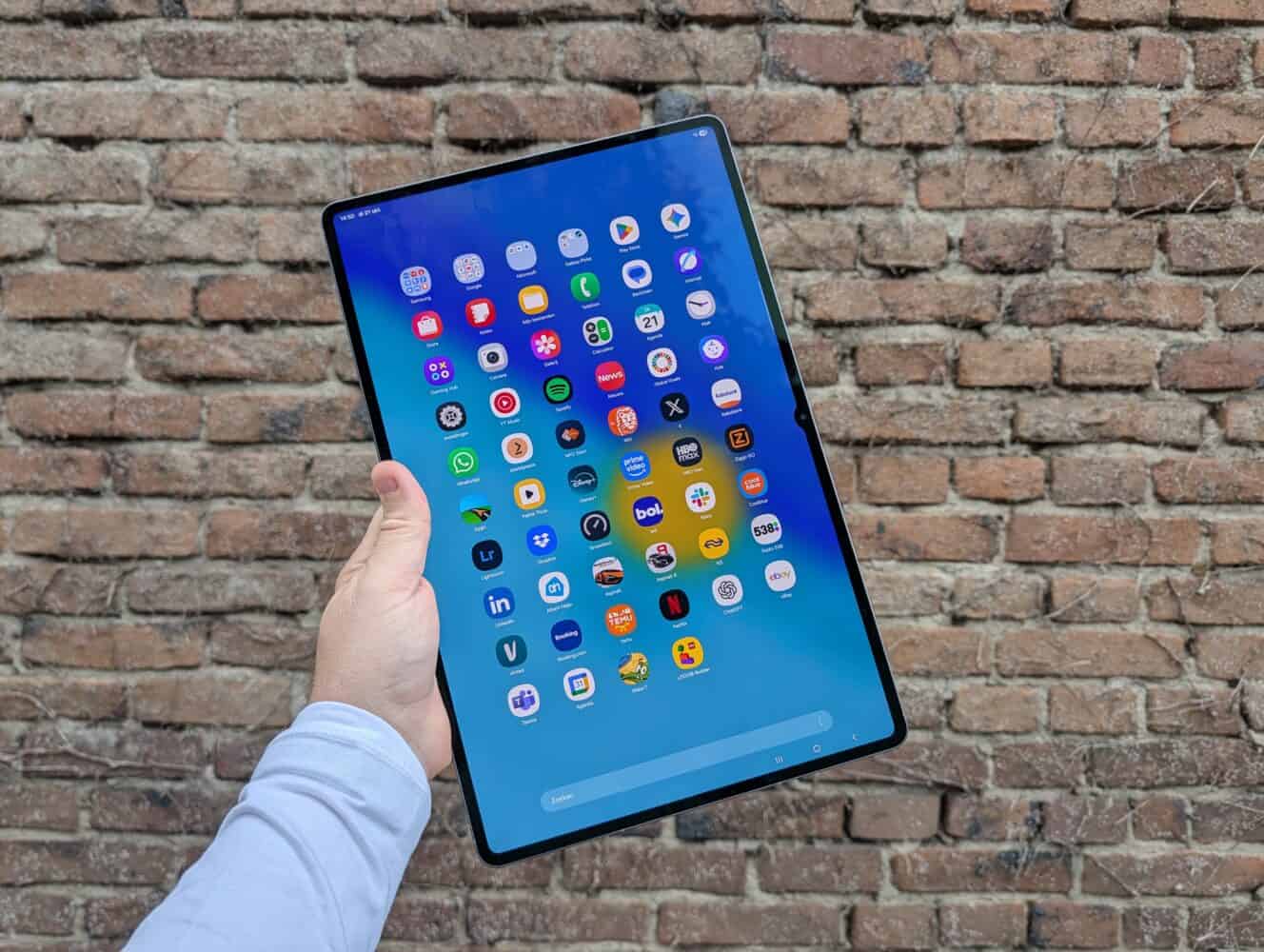 Een hand houdt een grote Samsung Galaxy Tab S11 Ultra vast die een raster van verschillende app-iconen weergeeft, met een bakstenen muur op de achtergrond.