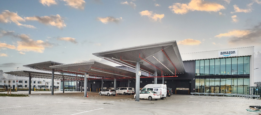 Sorteercentrum Amazon in Rozenburg Modern magazijn van Amazon met grote metalen luifels, verschillende geparkeerde witte bestelwagens en een glazen kantoorgedeelte onder een gedeeltelijk bewolkte hemel.