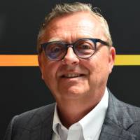 WD presenteert dunste hybride harde schijf ooit Een man van middelbare leeftijd met kort licht haar en een bril, draagt een geruit colbert en wit overhemd, staat voor een zwarte achtergrond met een oranje-gele horizontale streep.