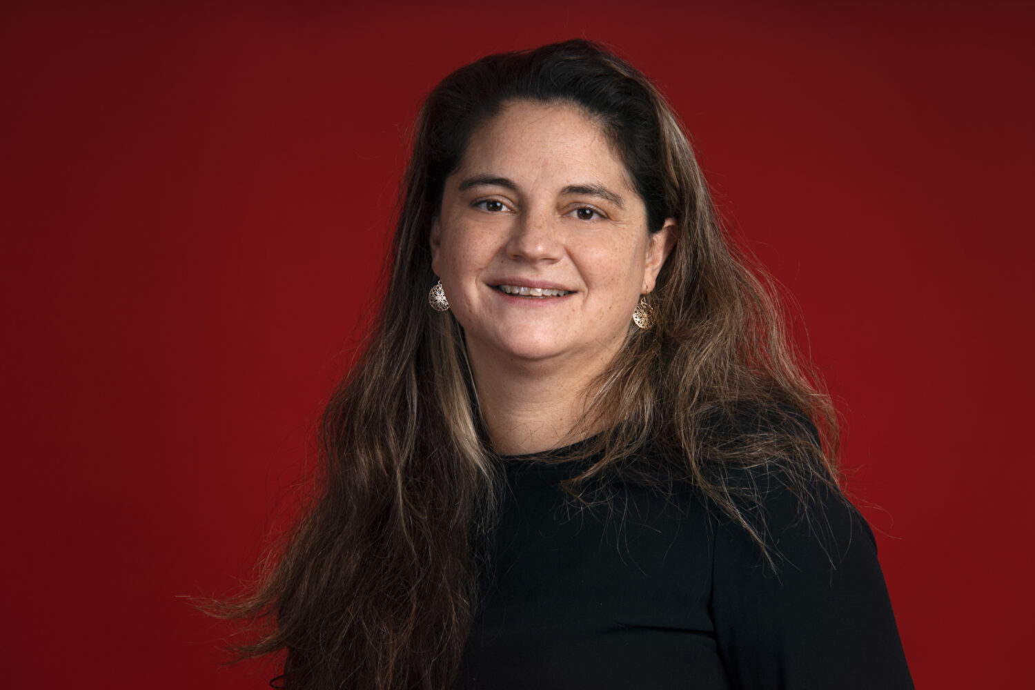 Een vrouw met lang bruin haar draagt een zwart topje en oorbellen poseert voor een effen rode achtergrond.