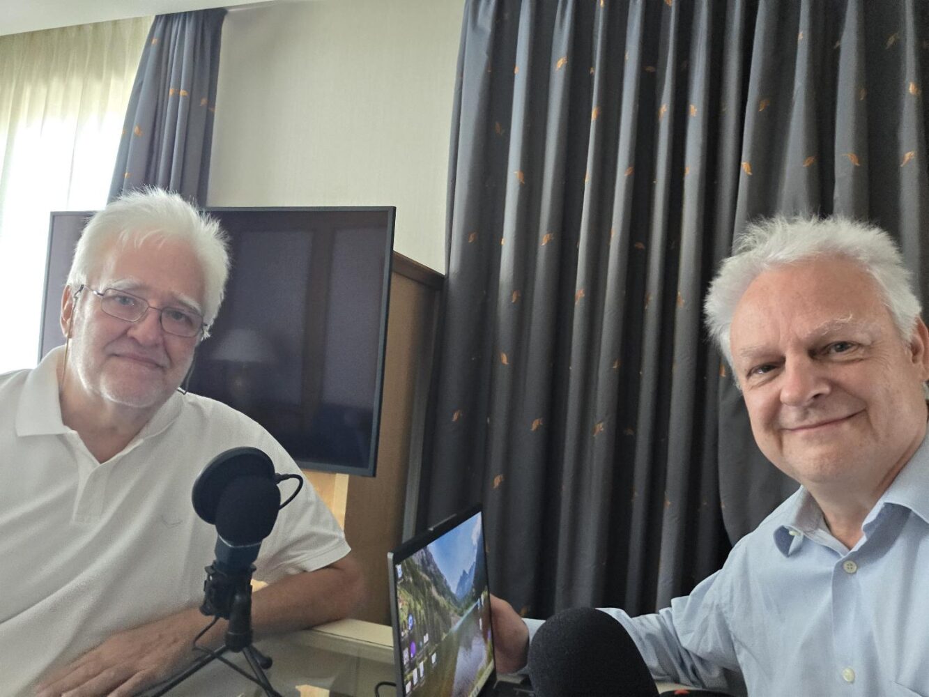 Twee oudere mannen met wit haar zitten aan een bureau met microfoons en een tablet, in een kamer met een tv en donkere gordijnen op de achtergrond.
