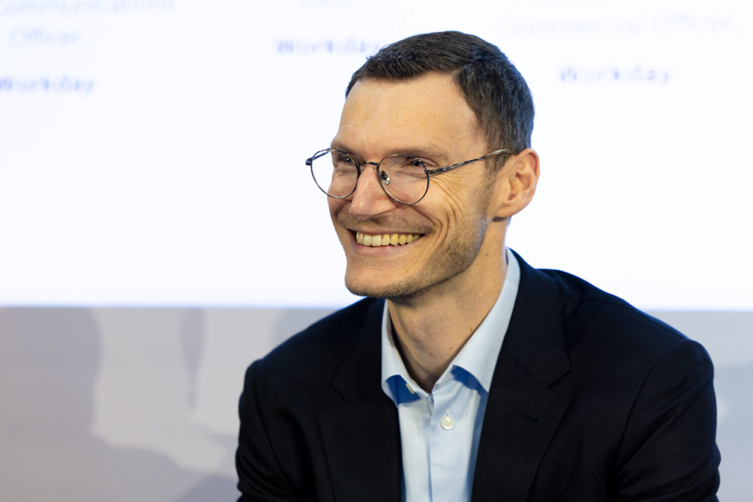 Een man met een bril en een donker colbert glimlacht terwijl hij binnen zit voor een wazige witte achtergrond, die het professionalisme van de Workday EU Sovereign Cloud-omgeving weerspiegelt.