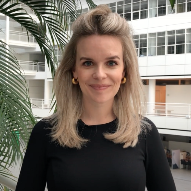 Een vrouw met schouderlang blond haar en gouden hoepeloorbellen staat binnen en draagt een zwart topje. Modern wit gebouw en groene plant op de achtergrond.