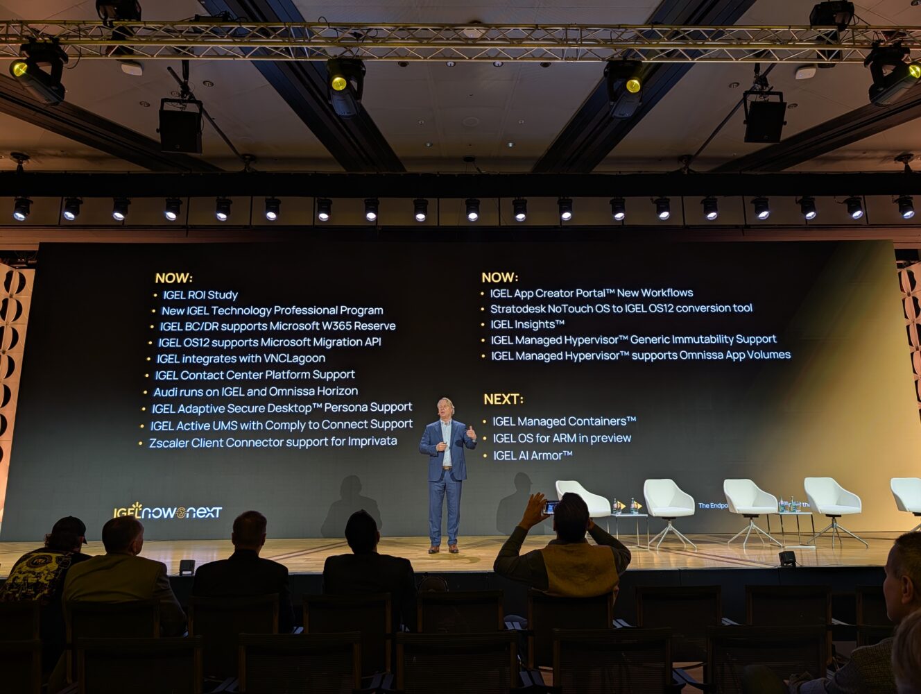 Een spreker staat op het podium van een technische conferentie voor een groot scherm met de nieuwste IGEL-productupdates en trekt zo de aandacht van de geëngageerde deelnemers.