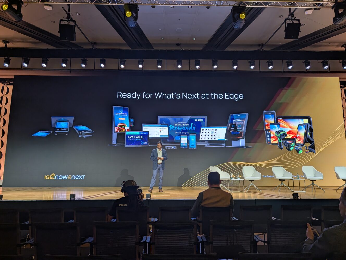 Een presentator staat op het podium voor een groot scherm met verschillende elektronische apparaten en de tekst "Ready for What's Next at the Edge," waarmee IGEL-oplossingen onder de aandacht worden gebracht op een technologie-evenement.