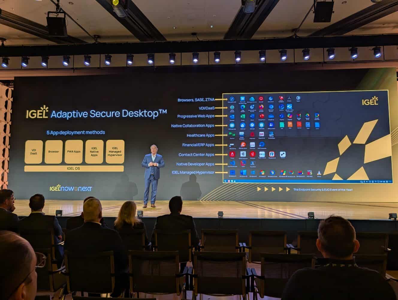 Een man staat op het podium en presenteert een slide over IGEL Adaptive Secure Desktop, met de IGEL desktop interface achter hem geprojecteerd aan een publiek in een vergaderzaal.
