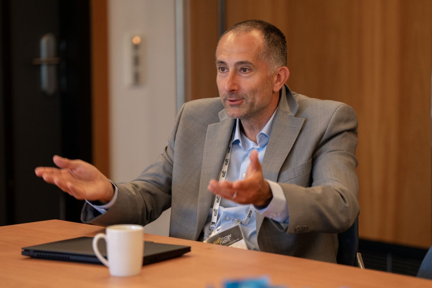 Nokia kraakt HTC af: ‘Innovatie is meer dan kleur’ Een man in een grijs pak zit aan een tafel met een IGEL laptop en een witte mok, en gebaart met open handen terwijl hij spreekt.