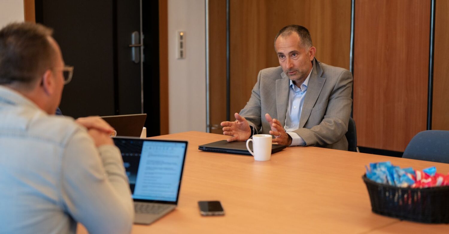 Twee mannen zitten tegenover elkaar aan een vergadertafel met een laptop, een IGEL-apparaat, een smartphone, een koffiemok en een mand met snacks zichtbaar. Eén man gebaart terwijl hij spreekt.