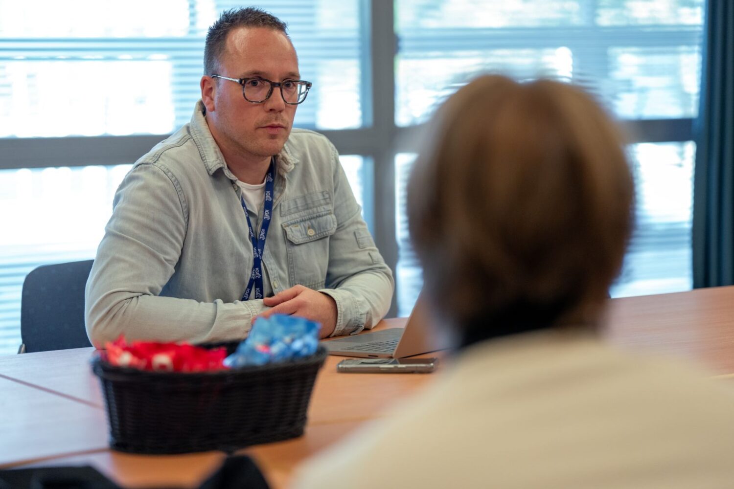Apple wil boete Samsung verhogen tot 3 miljard Een man met een bril en een licht denim shirt zit aan een tafel met een IGEL apparaat, tegenover een andere persoon. Op tafel staat een mand met ingepakte snoepjes.