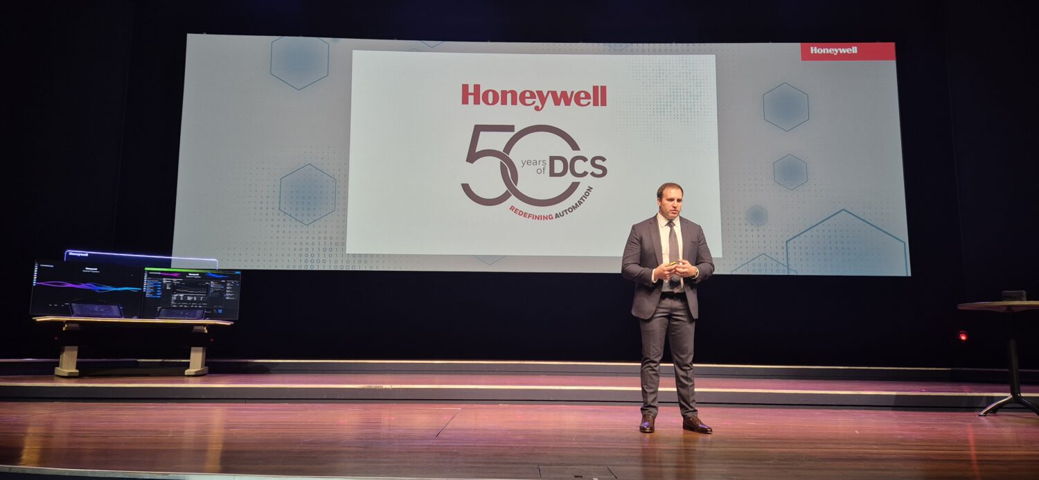 Een man in pak staat op het podium voor een groot scherm met daarop "Honeywell 50 years DCS" tijdens een presentatie-evenement.