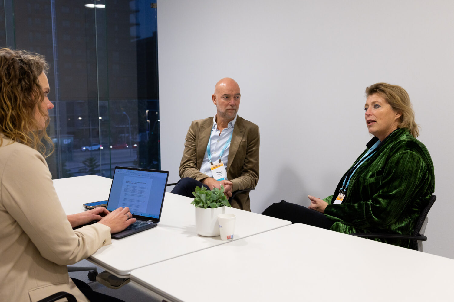 Drie mensen zitten rond een witte tafel in een kantoor; één typt op een laptop terwijl de andere twee een gesprek voeren. Een plantje en een kopje staan op tafel.