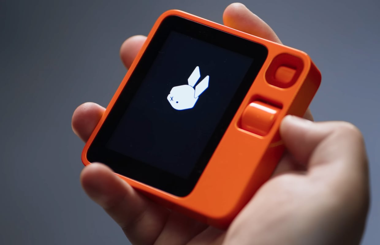 Een hand houdt een oranje handheld vast met een scherm waarop een wit konijn staat en twee oranje knoppen naast het scherm.
