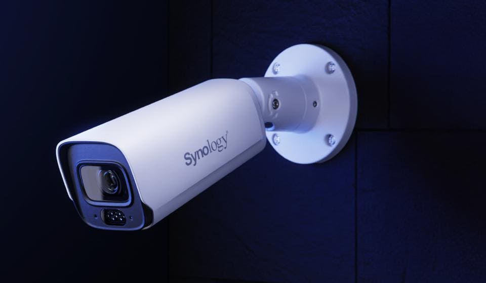 Een gestroomlijnde witte Synology beveiligingscamera is gemonteerd op een donkere muur, naar buiten gericht voor optimale bewaking.