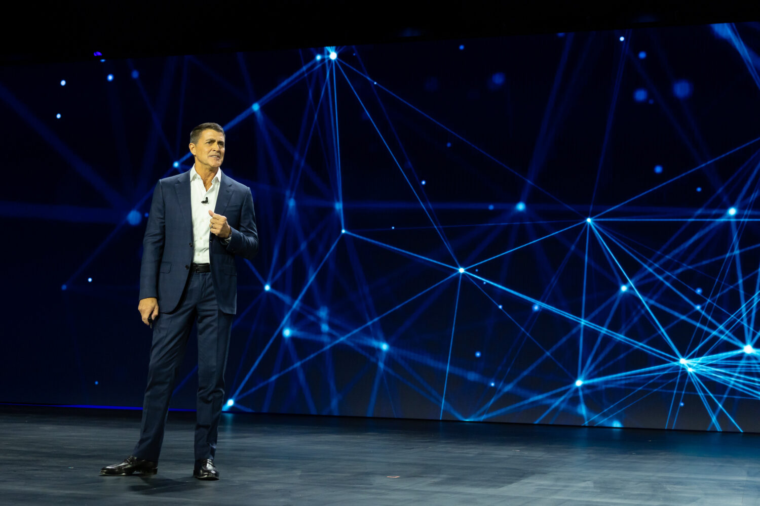 Een man in pak staat op het podium voor een groot scherm waarop blauwe onderling verbonden lijnen en knooppunten te zien zijn.