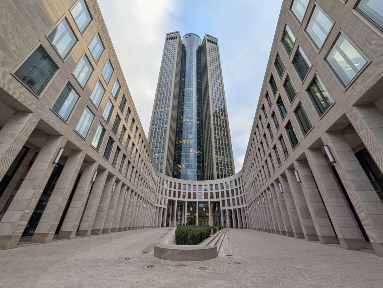 Symmetrisch uitzicht op een moderne glazen wolkenkrabber omlijst door twee gebogen stenen gebouwen, met een heldere hemel erboven en een stenen binnenplaats op de voorgrond - in verbluffend detail vastgelegd op de Google Pixel 10 Pro.