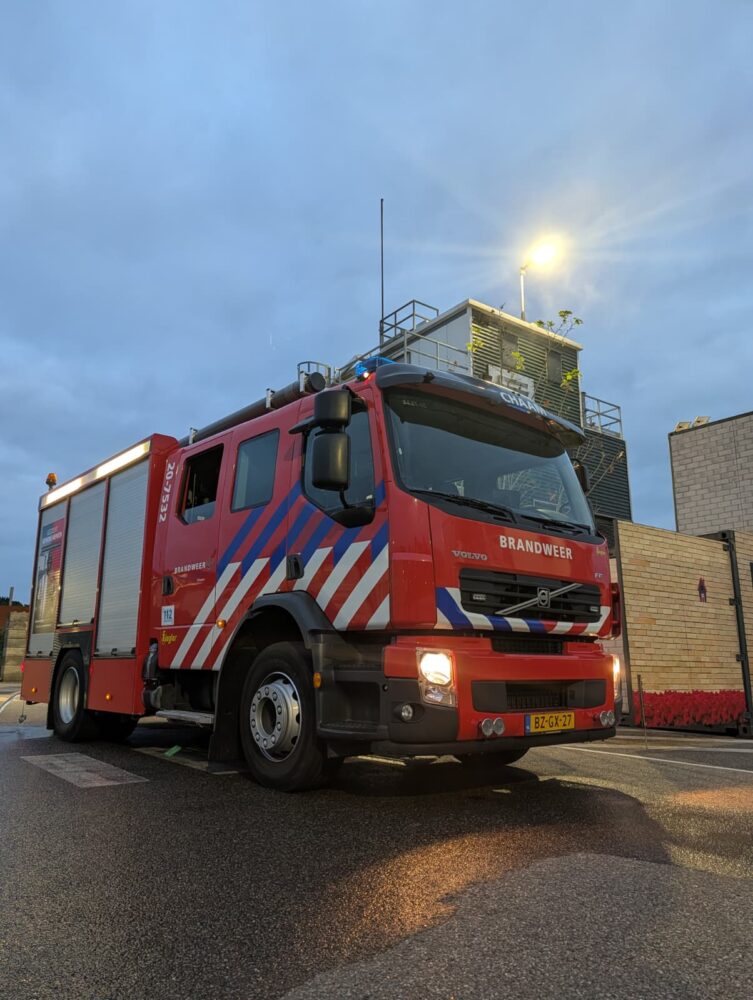 Een rode Nederlandse brandweerwagen met blauwe en witte strepen staat geparkeerd op een natte straat bij een modern gebouw in de schemering, in levendig detail vastgelegd met de Google Pixel 10 Pro.