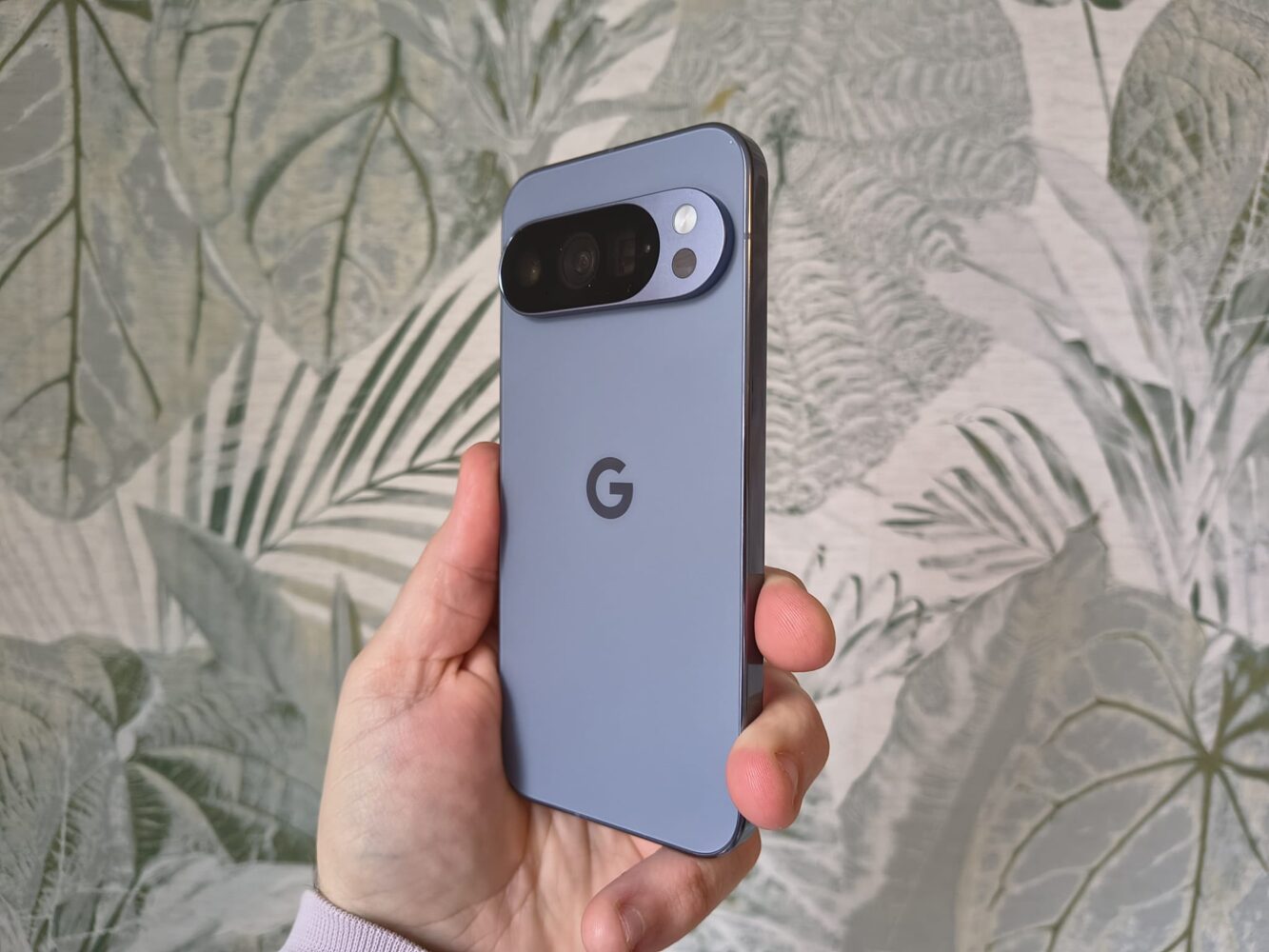 Een hand houdt een grijze Google Pixel 10 Pro smartphone met een horizontale camerabalk vast, voor een achtergrond met bladmotief.