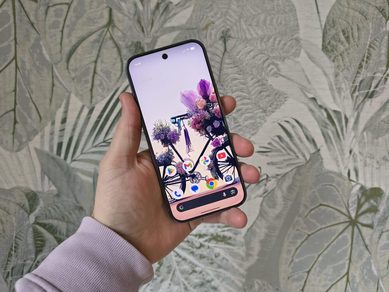 Een persoon met een Google Pixel 10 Pro met een achtergrond in de vorm van een bloemenfiets en verschillende app-icoontjes, tegen een achtergrond met bladmotief.