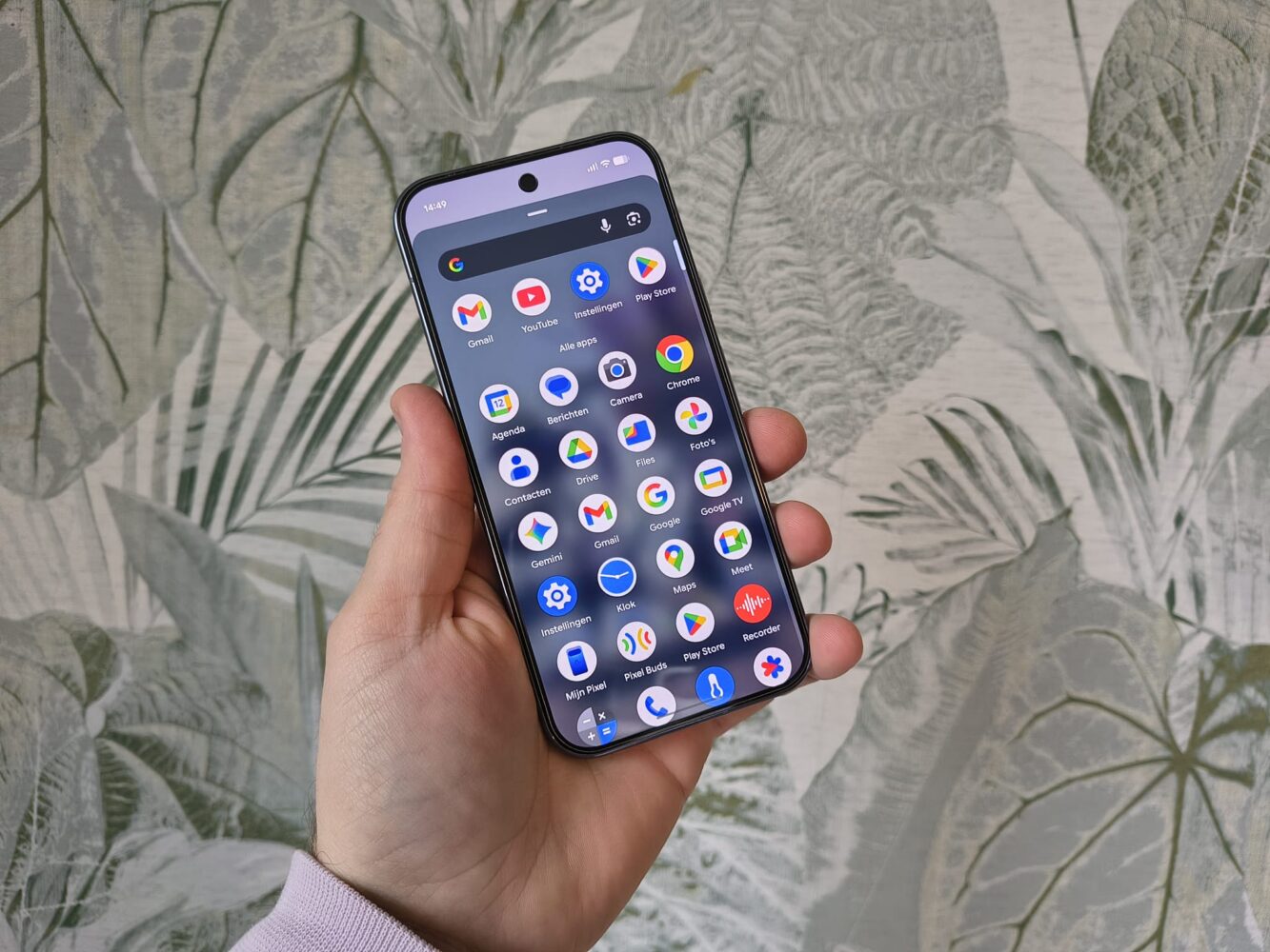 Een hand houdt een Google Pixel 10 Pro vast en toont de app-lade met verschillende Google-apps op het scherm; op de achtergrond is bladbehang zichtbaar.