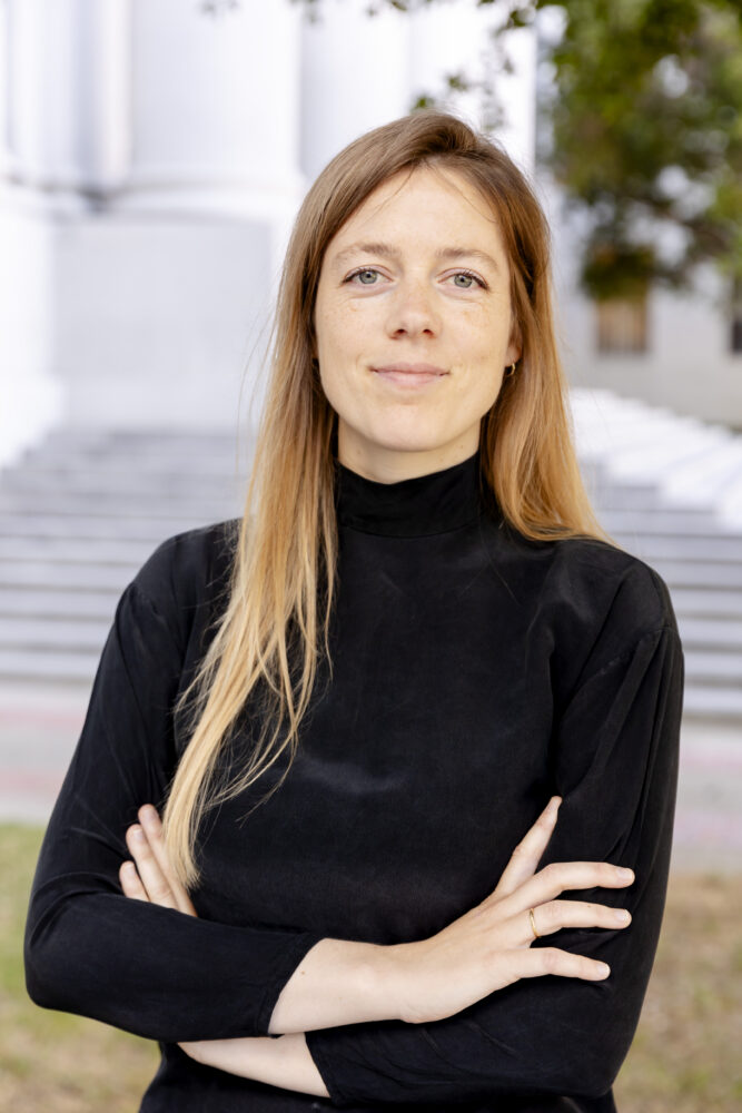 Een vrouw met lang blond haar en een zwart topje staat buiten met haar armen over elkaar en kijkt naar de camera. Op de achtergrond zijn trappen en bomen zichtbaar.