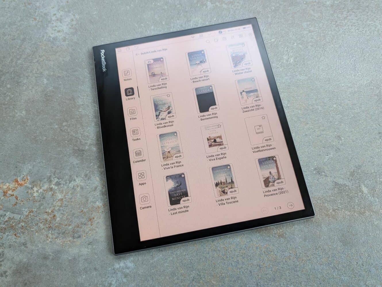 Een zwarte PocketBook InkPad Eo e-reader met een bibliotheek van digitale boekomslagen en bestandsmappen op een grijs oppervlak met structuur.