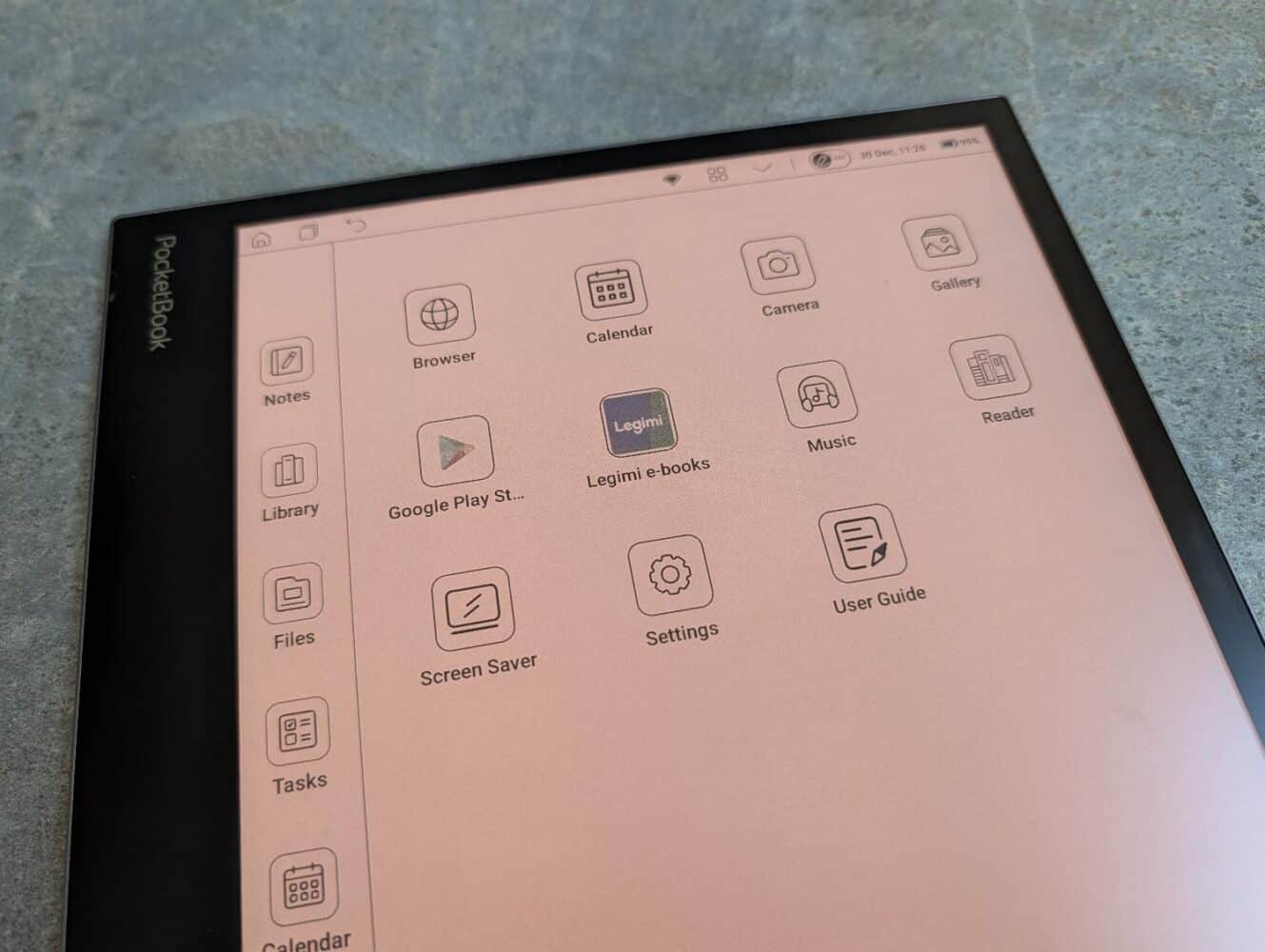 Een PocketBook InkPad Eo e-reader toont zijn startscherm met app-pictogrammen zoals Notities, Browser, Agenda, Camera, Galerij en meer op een grijs oppervlak.