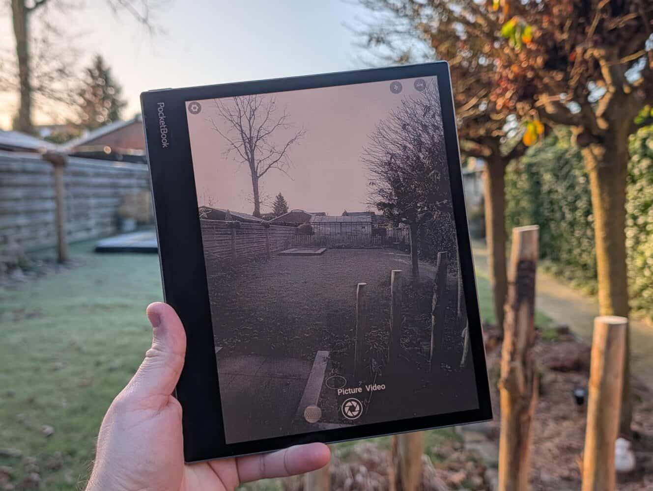 Een hand houdt een PocketBook InkPad Eo-tablet vast dat een zwart-witfoto weergeeft van dezelfde scène in de achtertuin die buiten op de achtergrond zichtbaar is.
