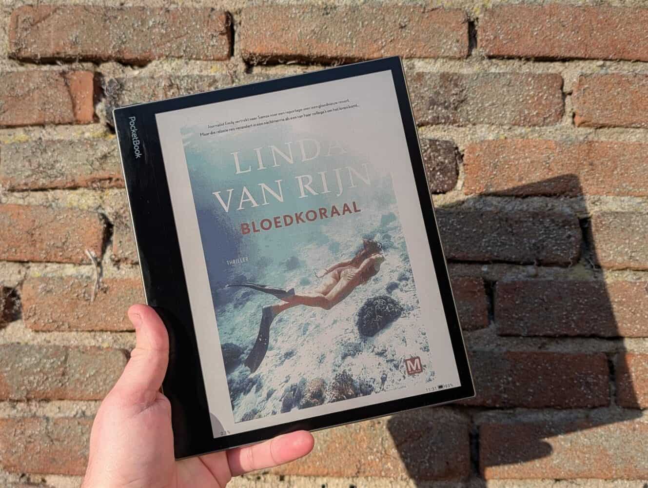 Een hand houdt een PocketBook InkPad Eo e-reader vast met daarop de cover van "Bloedkoraal" van Linda Van Rijn, tegen een bakstenen muur. Op de cover staat een duiker afgebeeld die onder water zwemt.