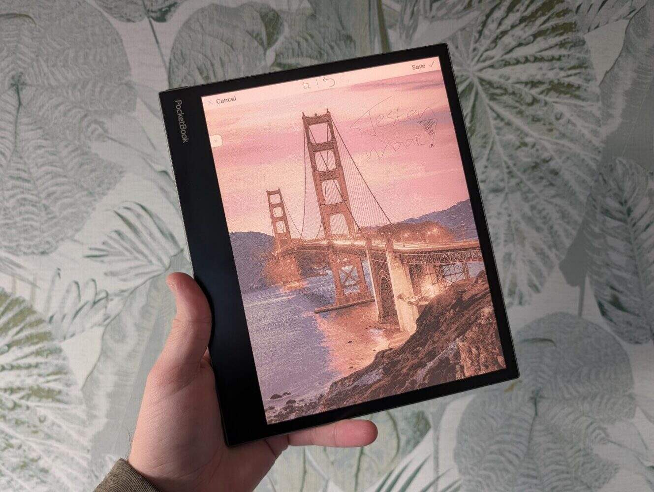 Een hand houdt een PocketBook InkPad Eo e-ink tablet vast dat een afbeelding van de Golden Gate Bridge toont met handgeschreven digitale notities op het scherm.