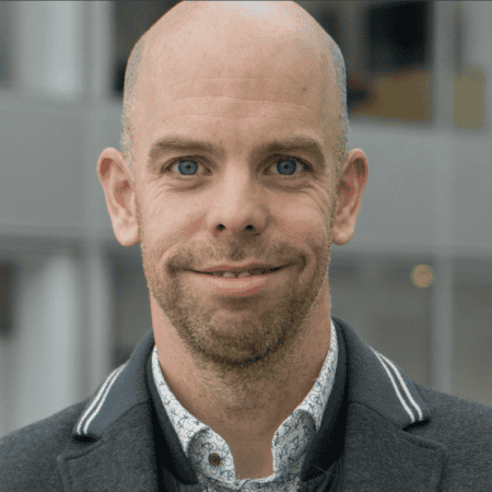 Een man met een kaal hoofd en licht gezichtshaar glimlacht lichtjes, draagt een shirt met patroon en een donker jasje, staat voor een wazige achtergrond van een gebouw.