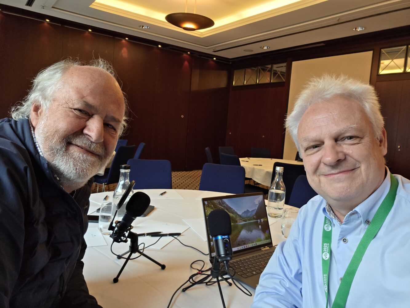 Twee mannen zitten aan een tafel met microfoons, waterflessen en een laptop in een vergaderzaal en bereiden zich voor op een opname of interview.