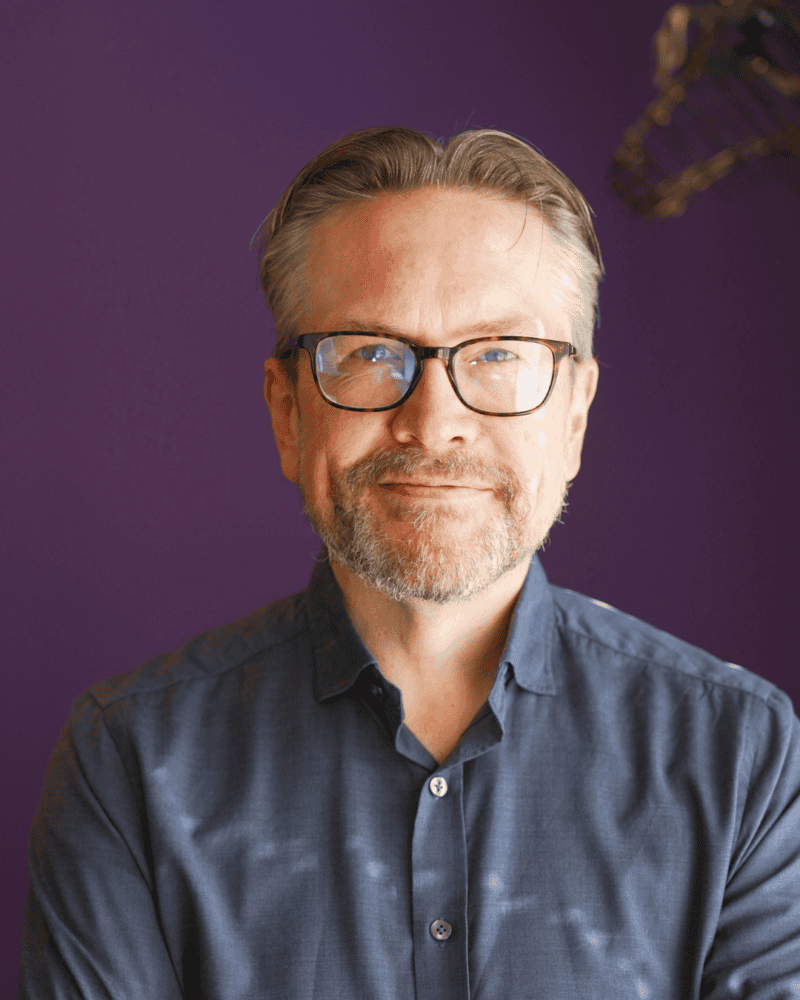 Een man van middelbare leeftijd met een bril en een korte baard glimlacht lichtjes, draagt een blauw overhemd en staat voor een effen paarse achtergrond.