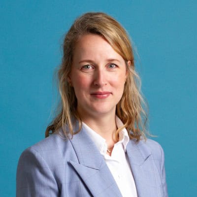 Een vrouw met blond haar draagt een lichtblauwe blazer en wit shirt voor een effen blauwe achtergrond.