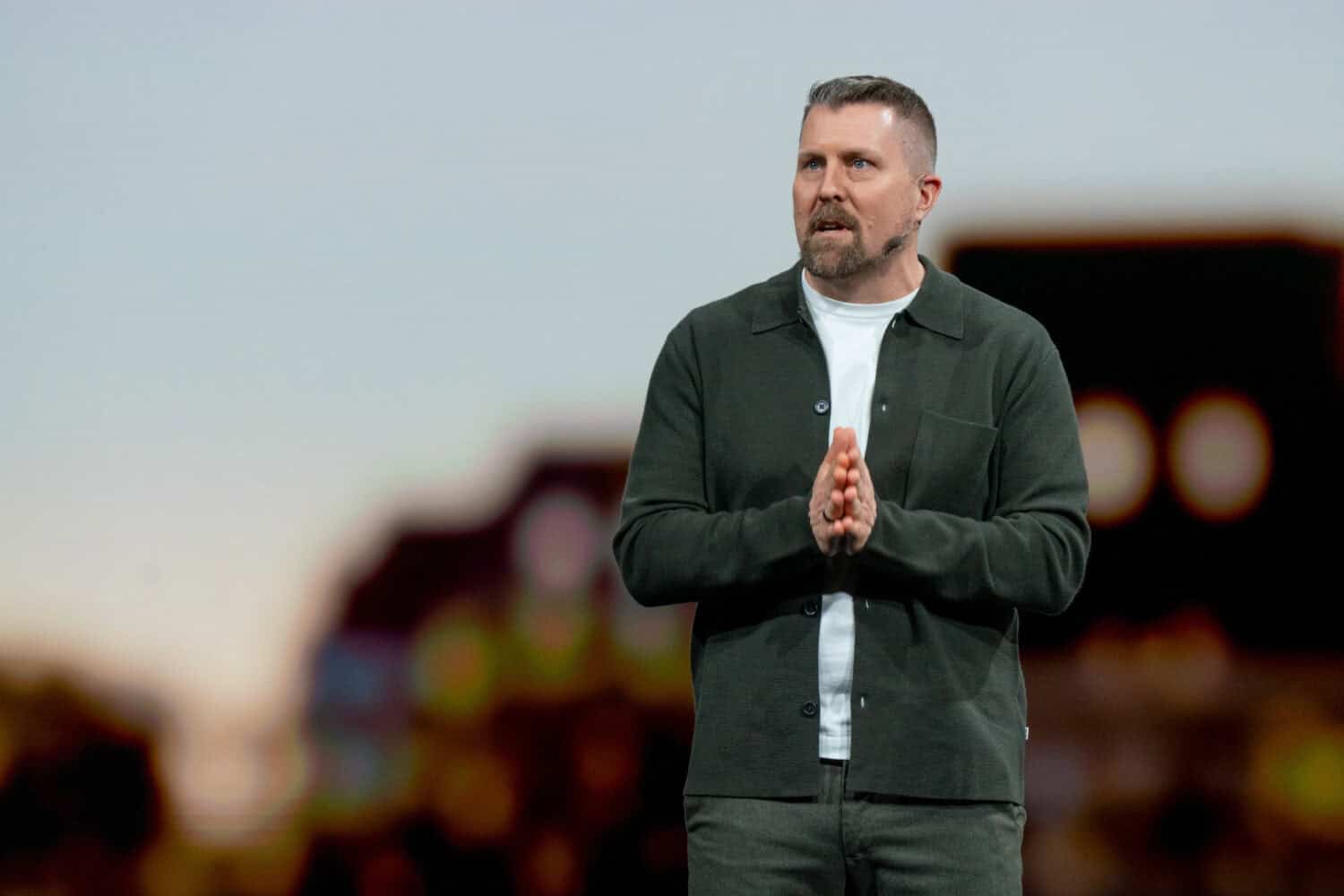 Een man met een koptelefoon op en een donkere jas staat op het podium met zijn handen in elkaar geklemd en spreekt tegen een wazige achtergrond.