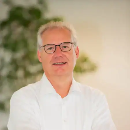 Een man van middelbare leeftijd met grijs haar en een bril, draagt een wit overhemd en staat binnen met een vage plant op de achtergrond.