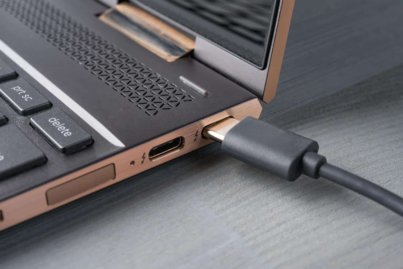 Een USB-C kabel is aangesloten op de oplaadpoort van een slanke, metalen laptop die op een grijs oppervlak rust.