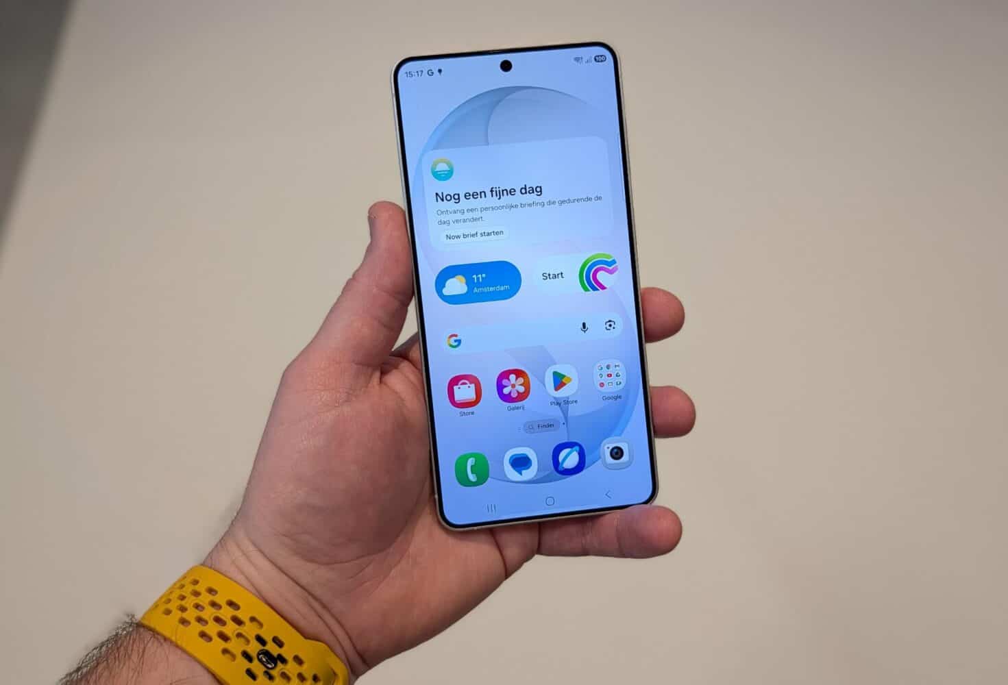Een hand die een Samsung Galaxy S26 vasthoudt toont een beginscherm met app-icoontjes en een weer-widget in het Nederlands. De persoon draagt een geel polsbandje.