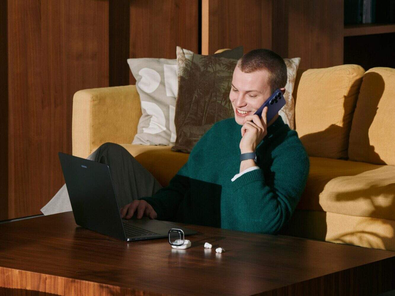Een persoon zit op de grond aan een koffietafel, gebruikt een laptop en praat op een Samsung Galaxy S26 smartphone, met draadloze oordopjes en een smartwatch op tafel en een gele bank op de achtergrond.