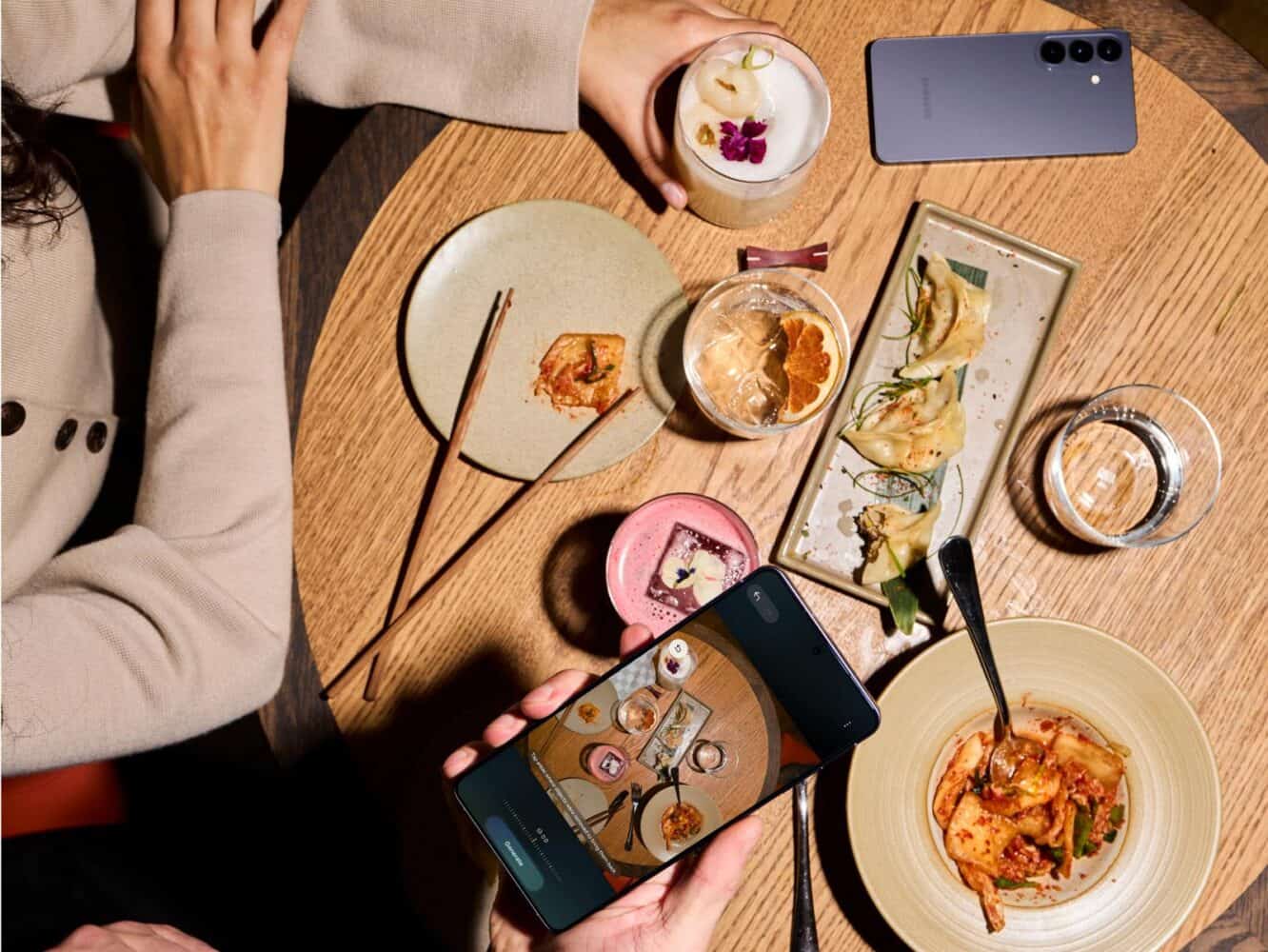 Twee mensen dineren aan een houten tafel met verschillende gerechten en drankjes; één persoon neemt een foto van het eten met zijn Samsung Galaxy S26 smartphone.