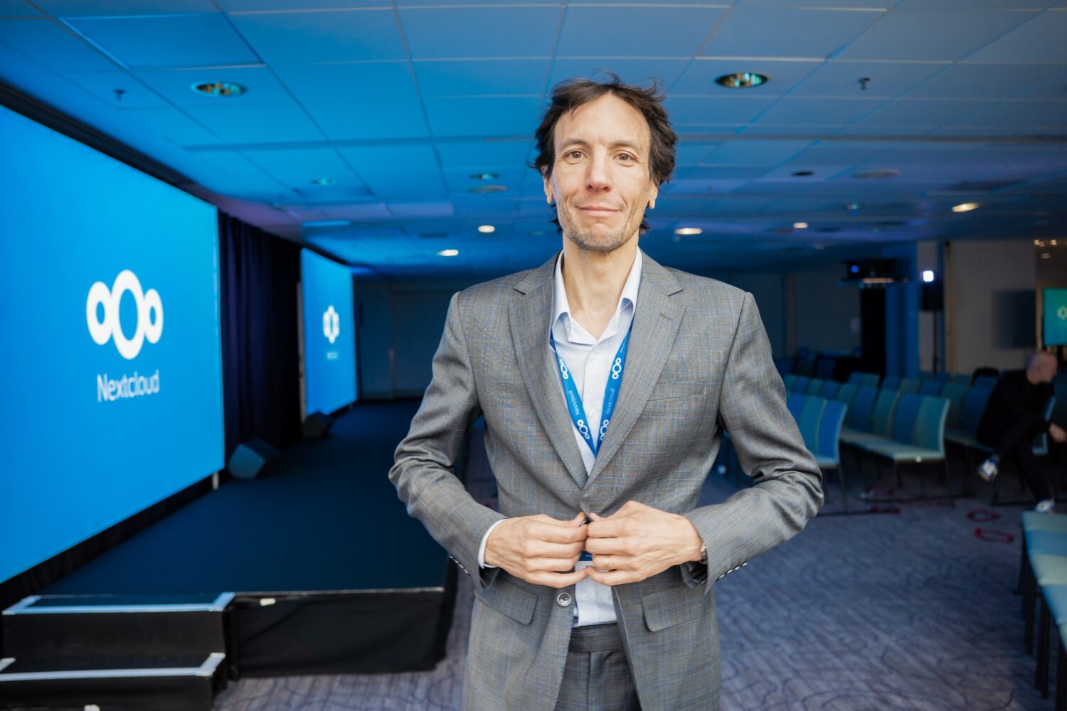 Een man in een grijs pak staat in een vergaderzaal met Nextcloud branding op schermen op de achtergrond, met de nadruk op open source migratie en digitale soevereiniteit.