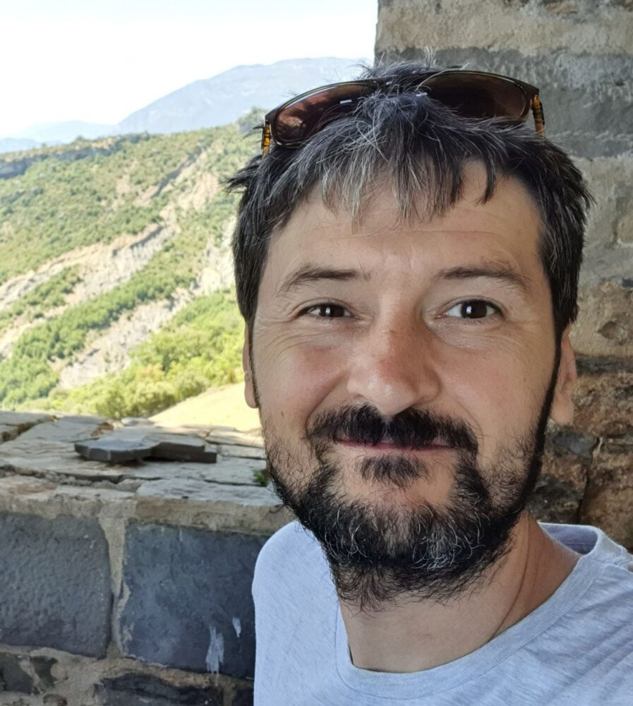 Een man met kort donker haar, een baard en een zonnebril op zijn hoofd lacht naar de camera met een schilderachtig berglandschap op de achtergrond, waarmee hij een moment van digitale soevereiniteit en avontuur vastlegt.