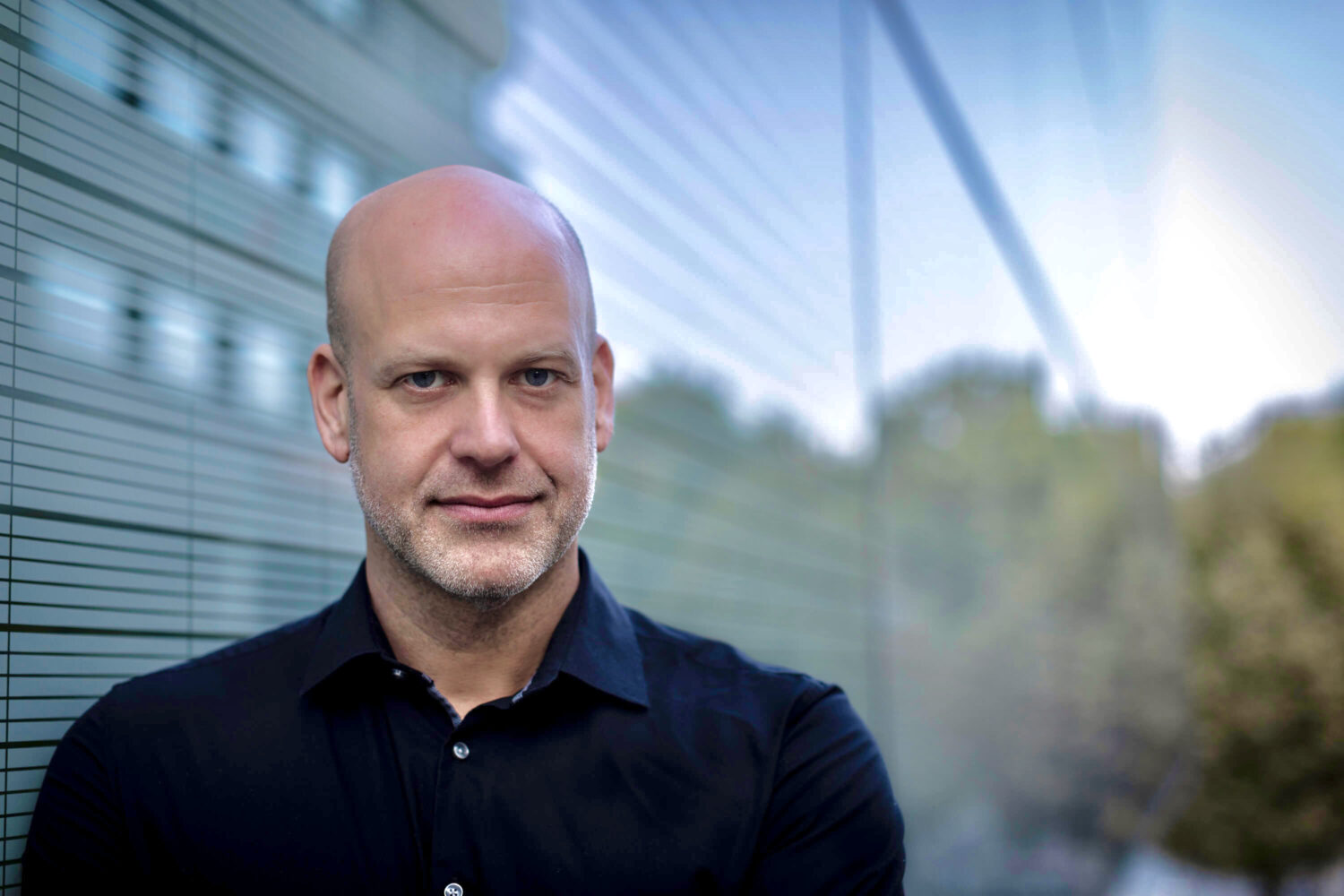 Een kale man in een zwart shirt staat voor een glazen wand met groen en een gebouw weerspiegeld in de achtergrond, symbool voor digitale soevereiniteit en de transparantie van open source migratie.