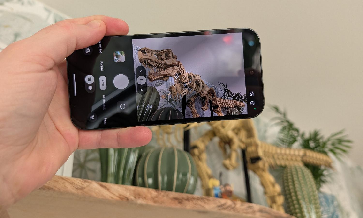 Een hand houdt een smartphone vast, uit een Google Pixel 10a review, en maakt een foto van een dinosaurus skelet op een plank, met planten en decor zichtbaar op de achtergrond.